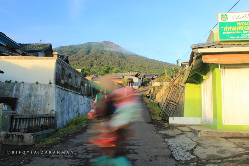 Warga dusun melintas di jalan kampung dengan latar belakang Gunung Slamet